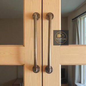 May include: Two brass door handles mounted on light wood panels. Each handle is a long, curved tube attached to circular plates with screws. The handles have a vintage, industrial aesthetic. The TukuBrass logo is visible.
