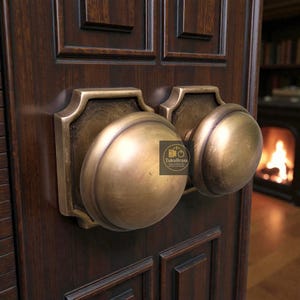 Könnte beinhalten: Nahaufnahme von antiker Messing-Türbeschlägen mit zwei großen, runden Türknöpfen. Die Knöpfe sind auf rechteckigen Rückplatten mit dekorativen Kanten montiert. Die Tür ist aus dunklem Holz mit eingelassenen Paneelen gefertigt. Die Beschläge haben ein Vintage-Aussehen.