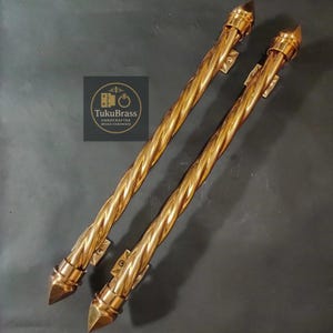 Puede incluir: Dos manijas de puerta de metal dorado retorcido con extremos puntiagudos. Las manijas tienen un diseño en espiral y están montadas en pequeñas placas rectangulares. El logotipo "TukuBrass HANDCRAFTED BRASS HARDWARE" es visible.