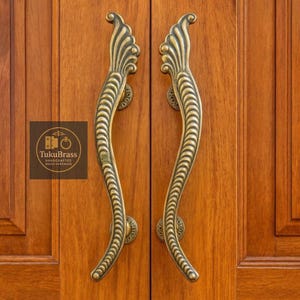 May include: A pair of ornate brass door handles with a curved design and decorative detailing. The handles are mounted on a wooden door, showcasing the hardware's elegant form and craftsmanship. The logo "TukuBrass Handcrafted Brass Hardware" is visible.