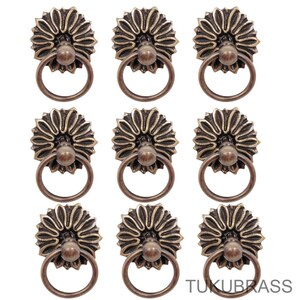 May include: Nine antique bronze cabinet pulls with a floral design. Each pull features a round ring handle and a decorative rosette base. The pulls are arranged in a grid pattern against a white background. The text "TUKUBRASS" is visible in the bottom right corner.