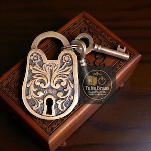 May include: An antique brass padlock and key set. The padlock has an ornate floral design. The key is inserted in the keyhole. The lock and key are resting on a wooden box with carved details. The brass has a warm, aged finish.
