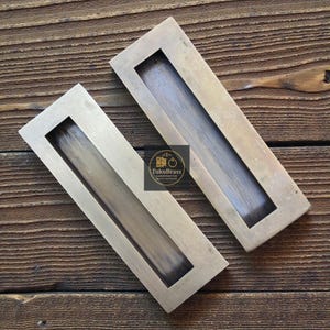 May include: Two rectangular brass recessed pulls with a dark wood grain interior. The pulls are set on a wooden surface. The logo "TukuBrass HANDCRAFTED BRASS HARDWARE" is visible.