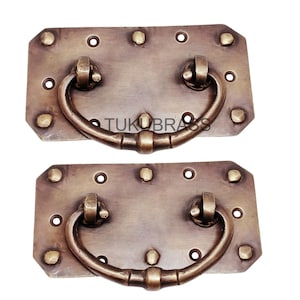 May include: Two antique brass drawer pulls with a rectangular backplate and a curved handle. Each pull has decorative rivets and the text "TUKUBRASS". These vintage-style pulls are ideal for furniture restoration or adding a classic touch to cabinets.