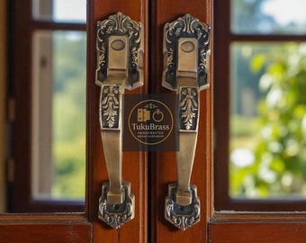 May include: Two antique brass door handles with ornate floral and scrollwork designs. The handles are mounted on a dark wooden door. The image includes the text "TukuBrass Handcrafted Brass Hardware".