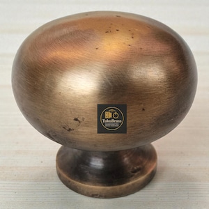 Może przedstawiać: Brązowy, kulisty uchwyt do szafki z zaokrągloną podstawą. Gałka ma szczotkowane metalowe wykończenie i logo z napisem "TukuBrass Handcrafted Brass Hardware". Okucia są prawdopodobnie do mebli.