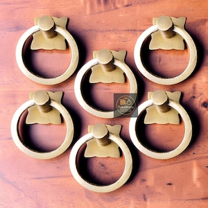 May include: Six brass ring pulls with a circular handle and a decorative backplate. The pulls are arranged on a wooden surface. The ring pulls are a warm gold color.