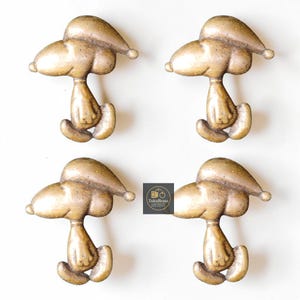 May include: Four bronze-colored Snoopy-shaped cabinet knobs. Each knob features Snoopy's iconic form, with a large head, floppy ears, and a seated pose. The knobs are set against a white background, with a logo in the lower right corner.