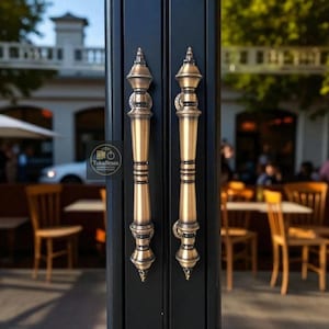 May include: Two ornate brass door handles with a dark background. The handles have a detailed design with a conical top and bottom, and a cylindrical body. The handles are mounted on a black door. The background is blurred, showing a restaurant setting.