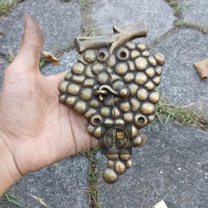 May include: A bronze-colored metal clasp shaped like a bunch of grapes, with a branch at the top. The clasp has a hook closure and is held in a hand. The product has the text "TukuBrass" on a sticker.