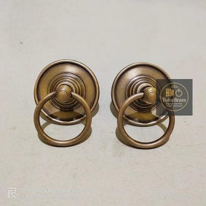 May include: A pair of antique brass cabinet pulls with ring handles. Each pull features a circular backplate and a tiered design. The ring handles are attached to the centre of the backplate. The pulls are made of brass and have a vintage finish.