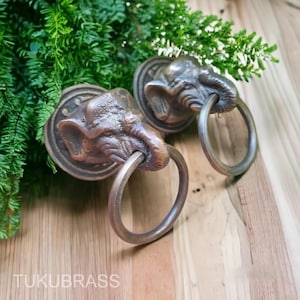 May include: Two bronze elephant head door knockers with circular handles. The elephant heads are detailed with textured skin and tusks. The knockers are mounted on circular backplates. The word "TUKUBRASS" is printed at the bottom of the image.