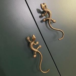 Puede incluir: Dos tiradores de armario con forma de lagarto de color bronce. Los tiradores tienen piel texturizada y colas curvas. Están montados en una puerta de armario verde oscuro con una costura diagonal.