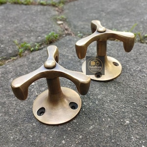 May include: Two antique brass coat hooks with a three-pronged design. Each hook has a circular base with three screw holes. The hooks are made of a warm, golden-toned brass and are set against a textured grey surface.