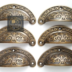 May include: Six antique brass drawer pulls with an ornate, arched design. Each pull features a central decorative element and intricate detailing along the edges. The pulls are a warm, aged brass color, suggesting vintage or rustic decor.