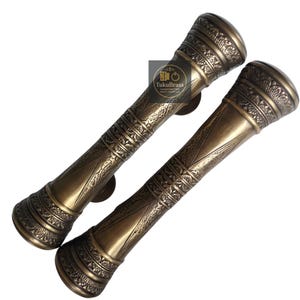 May include: Two antique bronze door handles with intricate, detailed patterns. The handles have a cylindrical shape with decorative bands and a circular end. The logo "TukuBrass" is visible on the image.