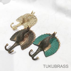 May include: Three decorative brass wall hooks shaped like stingrays. One is gold, one bronze, and one has a green base. Each hook features a curved tail and a detailed, textured design. The text "TUKUBRASS" is visible.