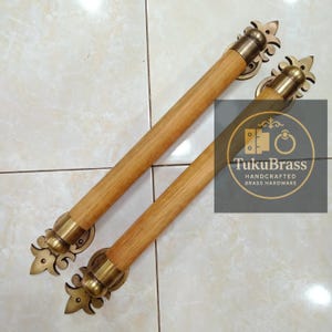 May include: Two wooden and brass door handles. The handles have a light brown wooden center with brass accents and decorative finials. The handles are handcrafted brass hardware, as indicated by the text "TukuBrass".