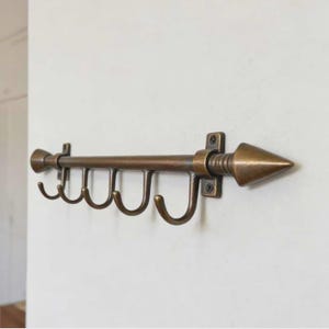 Puede incluir: Un perchero de pared de color bronce con cinco ganchos curvos. El perchero tiene una barra horizontal con un extremo decorativo en forma de flecha. Los ganchos están espaciados uniformemente a lo largo de la barra, y el perchero está montado en una pared blanca.