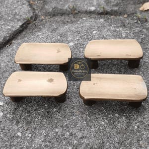 Peut inclure: Quatre petits tabourets rectangulaires en bois aux coins arrondis. Les tabourets ont un dessus brun clair et des pieds brun foncé. Les tabourets sont disposés sur une surface grise et texturée. Le logo "TukuBrass" est visible.