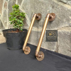 May include: Two bronze-colored pipe handles with flange mounts. The handles are angled and connected with elbow joints. The handles are mounted on a stone wall. The logo "TukuBrass HANDCRAFTED BRASS HARDWARE" is visible.