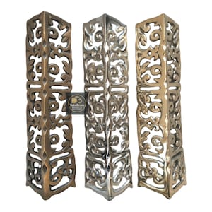 May include: Three decorative metal corner brackets with detailed openwork patterns. The brackets are in bronze, silver, and gold hues. The "TukuBrass" logo is visible on one of the brackets.