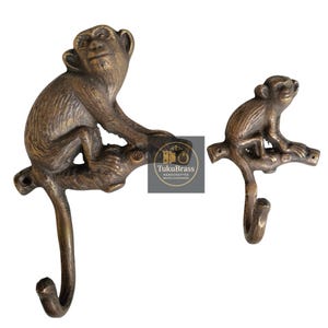 May include: Two brass monkey-shaped hooks. The larger monkey sits on a branch, with a hook extending downward. The smaller monkey also sits on a branch with a hook. The hooks are a bronze color.