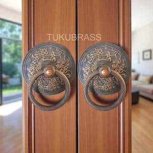 May include: Two antique bronze door handles with a circular design and ring pulls. The handles are mounted on a wooden door. The text "TUKUBRASS" is visible on the door.
