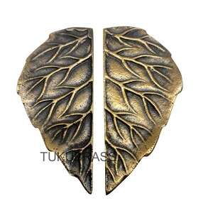 Może przedstawiać: Dwa uchwyty do drzwi w kształcie liścia w kolorze brązu. Każdy uchwyt ma szczegółowe żyłkowanie i teksturowaną powierzchnię. Uchwyty są wykonane z metalu i przeznaczone do montażu na drzwiach. Tekst "TUKU BRASS" jest widoczny w lewym dolnym rogu obrazu.