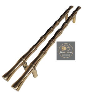 May include: Two brass-colored bamboo-style door pulls. The pulls are designed to resemble bamboo stalks, with segmented sections and a curved handle. The TukuBrass logo is in the lower right corner.