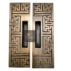 May include: Two bronze-colored door pulls with a geometric maze-like design. Each pull has a rectangular shape with a central, recessed handle. The pulls are made of metal and are likely for a door or cabinet.