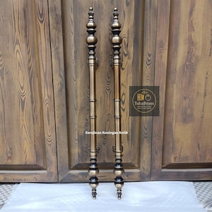May include: Two antique brass door handles with ornate detailing. The handles are vertical, with a dark brown and gold finish. The handles are mounted on a wooden door. The text "Kerajinan Kuningan Antik" is visible.