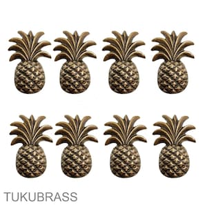 May include: Eight bronze pineapple-shaped cabinet knobs arranged in two rows of four. Each knob has detailed textures and a tropical design. The brand name "TUKUBRASS" is printed at the bottom.