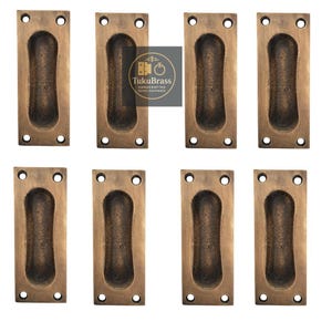 May include: Eight bronze-coloured recessed pull handles. Each rectangular handle has a rounded, oval-shaped indentation for gripping and four screw holes. The handles are arranged in two rows of four against a white background. The logo "TukuBrass" is visible.