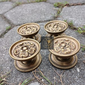 Może przedstawiać: Cztery mosiężne gałki do szafek w stylu antycznym z kwiatowym wzorem. Każda gałka ma okrągły, wypukły kwiatowy środek i dekoracyjną podstawę. Gałki są ułożone na szarej kamiennej powierzchni, prezentując ich styl vintage.