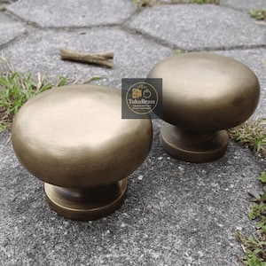 May include: Two antique brass cabinet knobs with a rounded top and a pedestal base. The knobs have a warm, golden-brown finish and are displayed on a textured gray surface. The logo "TukuBrass" is visible.
