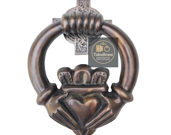 Könnte beinhalten: Bronzefarbener Türklopfer mit Claddagh-Design, einschließlich Herz, Krone und Händen, und einer kreuzförmigen Rückplatte. Der Klopfer hat einen Ringgriff und ist aus Metall gefertigt. Das Logo "TukuBrass Handcrafted Brass Hardware" ist sichtbar.