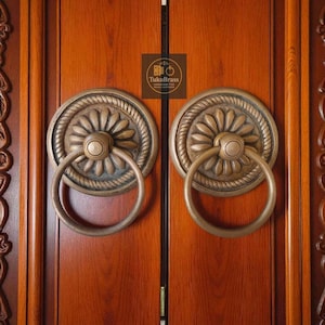 May include: Two antique brass door knockers with a floral design and ring handles, mounted on a wooden door. The door knockers are circular with a detailed rope-like border. The door is a warm brown color.