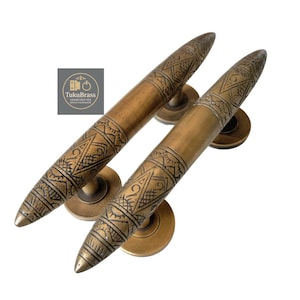 May include: Two antique brass door handles with intricate geometric patterns. The handles have a cylindrical shape with a pointed end and a circular base. The logo "TukuBrass HANDCRAFTED BRASS HARDWARE" is visible.