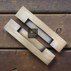 May include: Two rectangular brass cabinet pulls with a recessed handle area. The pulls have a brushed finish and are angled. The brand name "TukuBrass" is visible on a small square label.
