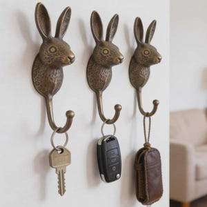 Puede incluir: Tres perchas de pared de color bronce con forma de cabeza de conejo. Cada gancho tiene una sección inferior curvada para colgar objetos. Una llave, un mando de coche y una funda de cuero cuelgan de los ganchos. Los ganchos están montados en una pared blanca.