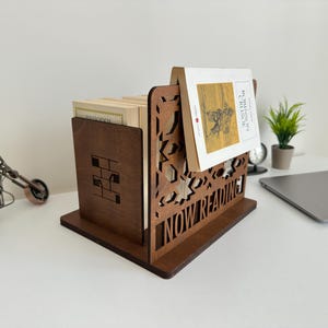 May include: A dark brown wooden book organiser. The organiser has a rectangular base and a vertical section with cut-out designs and the words "NOW READING". A book is displayed in the organiser. A small toy motorcycle is on the left.