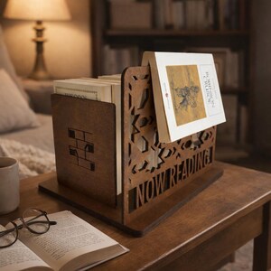May include: A wooden book holder with the words "NOW READING" cut into the front. The holder is on a wooden table with a cup of coffee, an open book, and glasses. A lamp and a bookshelf are in the background.