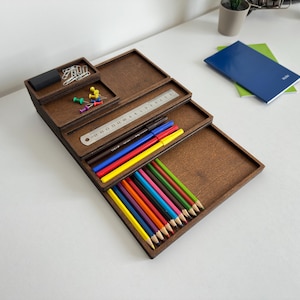 May include: A wooden desk organizer with multiple compartments. The organizer holds pencils, pens, push pins, paper clips, and a ruler. The pencils are various colors, including red, yellow, blue, and green. A blue notebook and a small potted plant are also visible.