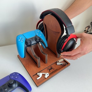 Puede incluir: Una estación de juego de madera con un mando azul y unos auriculares negros y rojos. La estación tiene ranuras para el mando y los auriculares, además de una pequeña bandeja con unidades USB y auriculares. Se ve un segundo mando morado.