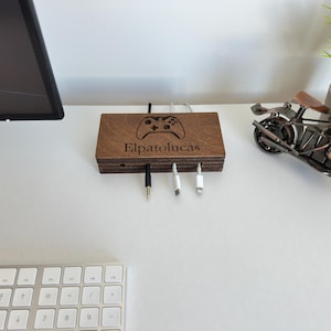 Puede incluir: Una estación de carga de madera con un diseño de mando de videojuegos y el texto "Elpatolucas". La estación tiene tres cables de carga y está sobre un escritorio blanco. También se ven un monitor de ordenador, un teclado y una moto decorativa.