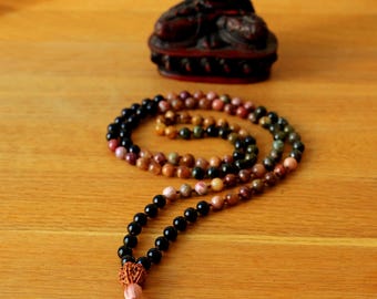 May include: A mala bead necklace with a pink tassel and a small Buddha statue. The necklace features black, brown, and pink beads. The Buddha statue is dark red and sits on a wooden surface.