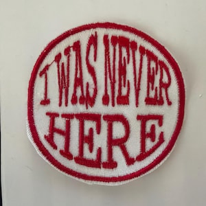 Może przedstawiać: Okrągła haftowana naszywka z białym tłem i grubą czerwoną obwódką. Napis "I WAS NEVER HERE" jest wyhaftowany czerwonymi wielkimi literami na środku naszywki, przy użyciu pogrubionej czcionki szeryfowej w całym projekcie.