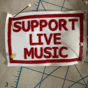 以下が含まれることがあります： 「SUPPORT LIVE MUSIC」と赤い刺繍が施された白い布パッチ。テキストは赤い縁で囲まれています。模様のある表面に、いくつかのピンでパッチが固定されています。