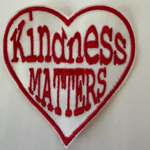 May include: A heart-shaped embroidered patch with a white background and a red border. The text "Kindness Matters" is stitched in red thread across the centre of the heart in a bold, stylised font, following the shape of the heart.